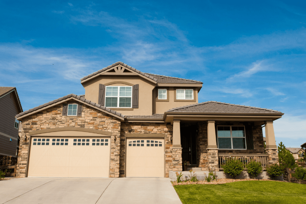 Front view of a two-story suburban house with a stone facade, beige garage doors, and a manicured lawn under a blue sky.